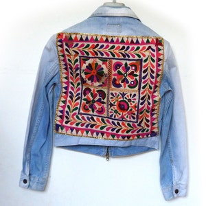 May include: Pale blue denim jacket with a vibrant embroidered panel on the back. The embroidery showcases floral and geometric motifs in pink, orange, green, and purple, edged with gold trim. The jacket has a zip and buttoned cuffs.