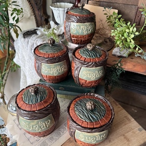 May include: A collection of ceramic sugar jars with lids. The jars are brown with a brick-like pattern and a green label that reads "Sugar". The lids are green and brown with a small knob on top. The jars are arranged on a wooden surface.