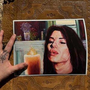 Puede incluir: Impresi&oacute;n de una mujer soplando un beso, con una vela encendida en primer plano. La obra de arte presenta una paleta de colores c&aacute;lidos, con un enfoque en el rostro de la mujer y la llama de la vela. Un jarr&oacute;n con flores es visible en el fondo.