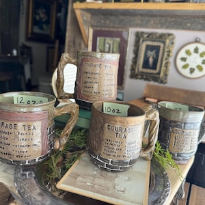 Könnte beinhalten: Vier Courage Tea-Tassen mit strukturierten, rustikalen Designs. Die Tassen haben ein braunes und beiges Farbschema mit ziegelartigen Details am Boden. Jede Tasse hat eine andere Kapazität, und der Text "Courage Tea" ist sichtbar.