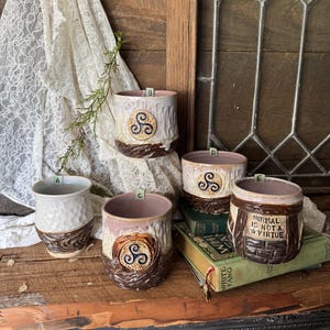 Pode incluir: Uma coleção de canecas de cerâmica artesanais com designs únicos. Cada caneca apresenta um exterior texturizado com uma base marrom e um emblema decorativo. Uma caneca tem o texto "NORMAL IS NOT A VIRTUE". As canecas são exibidas em uma superfície de madeira.