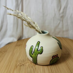 Könnte beinhalten: Eine kleine, runde Keramikvase mit cremefarbenem Boden und hellblauem Rand. Die Vase ist mit grünen Kaktusillustrationen verziert und enthält getrocknetes Ziergras. Die Vase steht auf einer Holzoberfläche.
