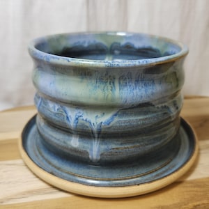 Puede incluir: Maceta de cerámica hecha a mano con platillo a juego. La maceta presenta un diseño acanalado y está esmaltada en tonos azules y verdes, con goteos blancos. El platillo es de color beige claro. Diámetro aproximado de 15 cm.