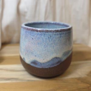 May include: A handmade ceramic tumbler with a rounded shape. The top half is a gradient of light blue and white, transitioning to a dark brown base. The rim has a dark brown accent. The tumbler sits on a wooden surface.