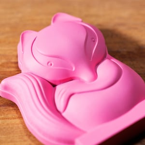 May include: A pink ceramic sculpture of a fox curled up in a sleeping position. The sculpture has a smooth, glossy finish and is detailed with a curled tail and a defined face.