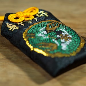 Puede incluir: Una pequeña bolsa de tela negra con un diseño bordado en oro y verde. La bolsa tiene un cordón amarillo atado alrededor.