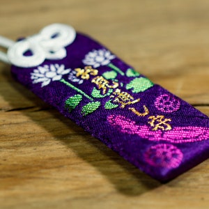 Puede incluir: Amuleto de tela morado con bordado dorado y blanco de flores y caracteres japoneses. El amuleto está atado con un cordón blanco.