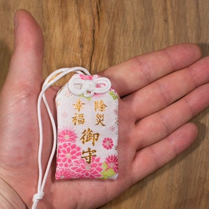 Puede incluir: Un amuleto de tela blanco y rosa con caracteres japoneses dorados que se leen "幸福 御守" (Amuleto de la felicidad). El amuleto tiene una cuerda blanca atada a &eacute;l y est&aacute; atado con un lazo blanco.
