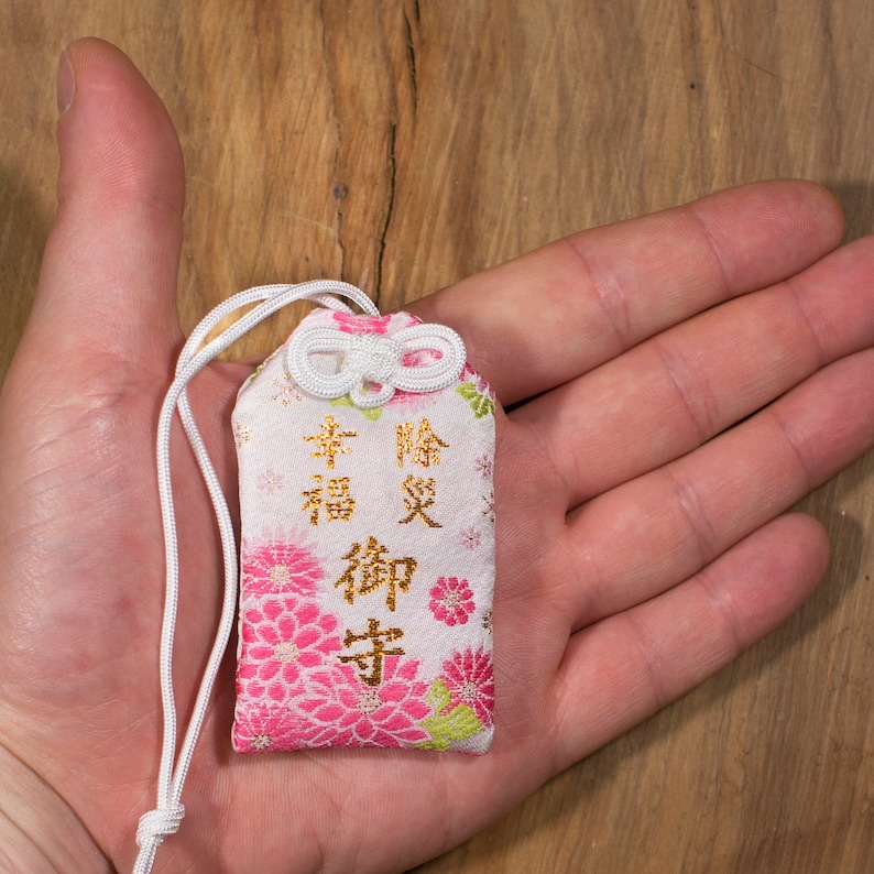 Puede incluir: Un amuleto de tela blanco y rosa con caracteres japoneses dorados que se leen "幸福 御守" (Amuleto de la felicidad). El amuleto tiene una cuerda blanca atada a &eacute;l y est&aacute; atado con un lazo blanco.