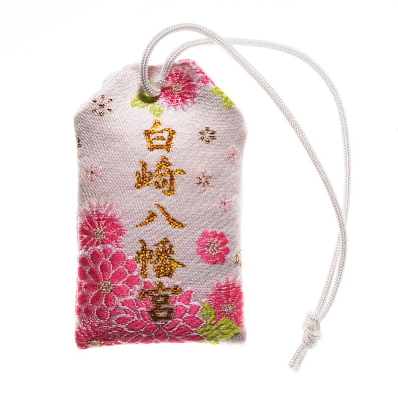 Puede incluir: Un amuleto de tela blanca con bordado floral rosa y caracteres japoneses dorados que dicen "白崎八幡宮". El amuleto est&aacute; unido a una cuerda blanca.