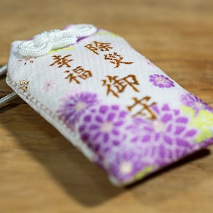 Puede incluir: Un amuleto de tela blanca con flores moradas y bordados dorados. El amuleto tiene los caracteres japoneses "幸福" (Felicidad) y "御守" (Amuleto) bordados en él.