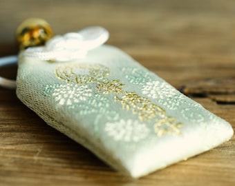 May include: A white fabric pouch with a gold and green floral pattern. The pouch has a white string tied around it and a small wooden bead.