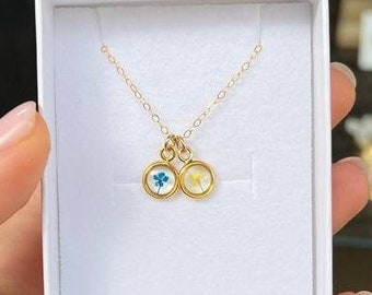 Könnte beinhalten: Eine goldene Kettenkette mit zwei kleinen runden Anhängern. Jeder Anhänger enthält eine gepresste Blume, eine blaue und eine gelbe.