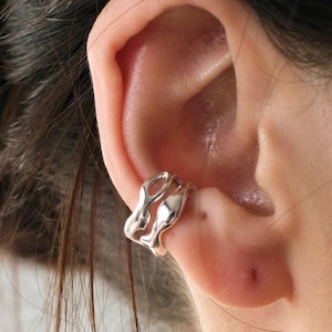 Puede incluir: Un ear cuff plateado con un diseño orgánico y fluido. El brazalete abraza el cartílago de la oreja, creando un look moderno y minimalista. La joya está hecha de un metal brillante y reflectante.