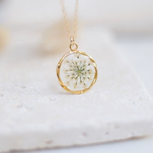 K&ouml;nnte beinhalten: Goldfarbene Halskette mit rundem Anh&auml;nger, der eine getrocknete wei&szlig;e Blume in klarem Harz enth&auml;lt. Die Halskette hat eine zarte Goldkette, die ein elegantes, von der Natur inspiriertes Accessoire schafft.
