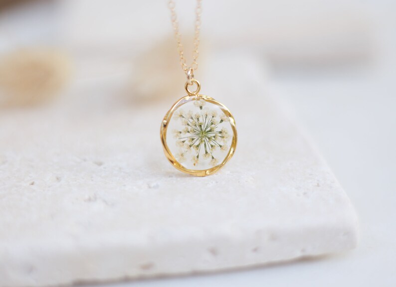 K&ouml;nnte beinhalten: Goldfarbene Halskette mit rundem Anh&auml;nger, der eine getrocknete wei&szlig;e Blume in klarem Harz enth&auml;lt. Die Halskette hat eine zarte Goldkette, die ein elegantes, von der Natur inspiriertes Accessoire schafft.