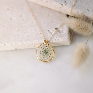 K&ouml;nnte beinhalten: Goldfarbene Halskette mit rundem Anh&auml;nger, der eine konservierte wei&szlig;e Blume in klarem Harz enth&auml;lt. Der Anh&auml;nger hat eine goldene L&uuml;nette und h&auml;ngt an einer feinen Goldkette. Der Hintergrund ist eine hellfarbige Steinoberfl&auml;che.