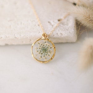K&ouml;nnte beinhalten: Eine goldfarbene Halskette mit einem runden Anh&auml;nger, der eine gepresste wei&szlig;e Blume enth&auml;lt. Der Anh&auml;nger ist in Gold gefasst und h&auml;ngt an einer feinen Goldkette. Der Hintergrund ist in einer weichen, neutralen Farbe gehalten, die den Schmuck hervorhebt.