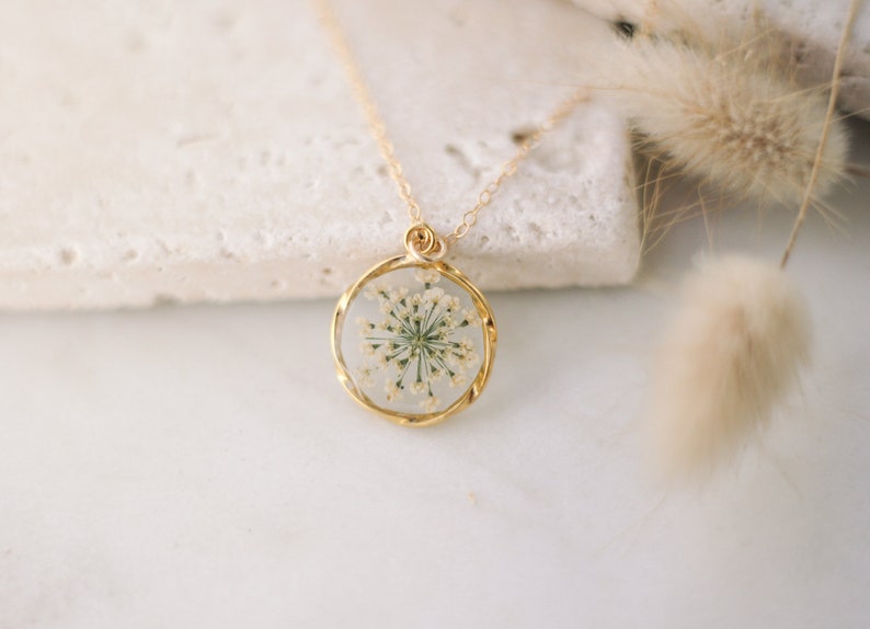 K&ouml;nnte beinhalten: Eine goldfarbene Halskette mit einem runden Anh&auml;nger, der eine gepresste wei&szlig;e Blume enth&auml;lt. Der Anh&auml;nger ist in Gold gefasst und h&auml;ngt an einer feinen Goldkette. Der Hintergrund ist in einer weichen, neutralen Farbe gehalten, die den Schmuck hervorhebt.