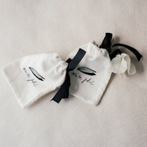 K&ouml;nnte beinhalten: Zwei kleine, cremefarbene Kordelzugbeutel mit schwarzen B&auml;ndern. Auf jeder Tasche befindet sich ein schwarz-wei&szlig;es Logo mit der Aufschrift "White petal". Neben einer der Taschen liegen ein paar wei&szlig;e Bl&uuml;tenbl&auml;tter.