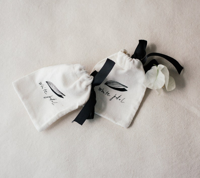 K&ouml;nnte beinhalten: Zwei kleine, cremefarbene Kordelzugbeutel mit schwarzen B&auml;ndern. Auf jeder Tasche befindet sich ein schwarz-wei&szlig;es Logo mit der Aufschrift "White petal". Neben einer der Taschen liegen ein paar wei&szlig;e Bl&uuml;tenbl&auml;tter.