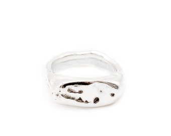 May include: A silver ring with a wide, irregular band featuring a textured, organic surface. The metal has a polished finish with small indentations and a slightly uneven, wavy edge, creating a distinct, non-uniform shape throughout the entire band.