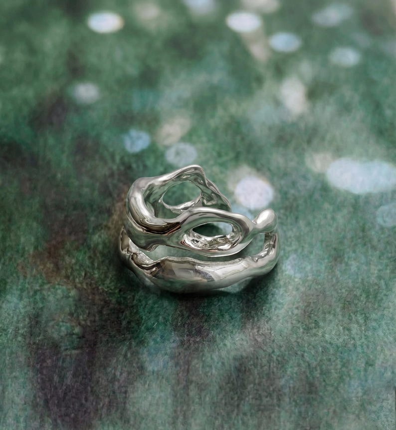 Pu&ograve; includere: Un anello d'argento dal design organico e fluido. L'anello presenta pi&ugrave; strati e spazi aperti, creando un aspetto unico e scultoreo. Il metallo lucido riflette la luce, evidenziando le curve e le trame dell'anello. L'anello &egrave; posto su uno sfondo verde sfocato.