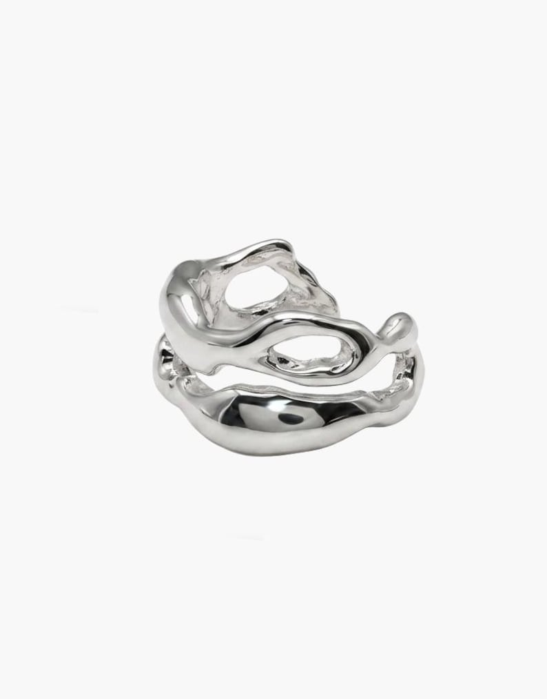 Pu&ograve; includere: Un anello d'argento dal design organico e fluido. L'anello presenta due fasce collegate con forme irregolari e fluide, creando un'estetica astratta e moderna. Il metallo lucido riflette la luce.