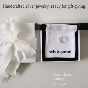 Pu&ograve; includere: Un orecchino a forma di petalo d'argento esposto in un cofanetto regalo bianco con bordo nero. La scatola contiene un sacchetto bianco con la scritta "white petal". A sinistra, un cofanetto regalo bianco con un nastro bianco. Testo: "Handcrafted silver jewelry, ready for gift-giving."