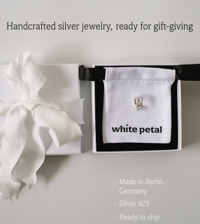 Pu&ograve; includere: Un orecchino a forma di petalo d'argento esposto in un cofanetto regalo bianco con bordo nero. La scatola contiene un sacchetto bianco con la scritta "white petal". A sinistra, un cofanetto regalo bianco con un nastro bianco. Testo: "Handcrafted silver jewelry, ready for gift-giving."