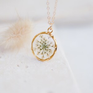 K&ouml;nnte beinhalten: Eine goldfarbene Halskette mit einem kreisf&ouml;rmigen Anh&auml;nger. Der Anh&auml;nger enth&auml;lt ein klares Harz mit einem zarten wei&szlig;en Blumenarrangement. Die Halskette ist goldfarben und der Anh&auml;nger hat einen Durchmesser von etwa 2,5 cm.