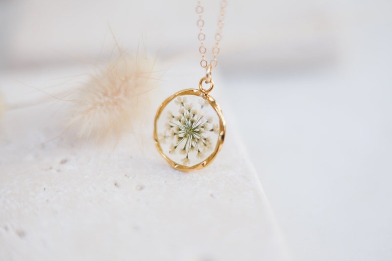 K&ouml;nnte beinhalten: Eine goldfarbene Halskette mit einem kreisf&ouml;rmigen Anh&auml;nger. Der Anh&auml;nger enth&auml;lt ein klares Harz mit einem zarten wei&szlig;en Blumenarrangement. Die Halskette ist goldfarben und der Anh&auml;nger hat einen Durchmesser von etwa 2,5 cm.