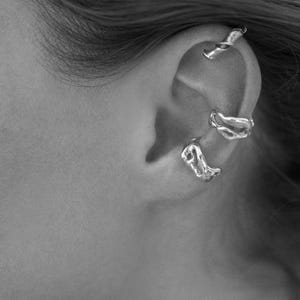 Pu&ograve; includere: Primo piano di un orecchio adornato da tre orecchini a polsino d'argento. Gli orecchini a polsino hanno un design organico e fluido, con un aspetto di metallo fuso strutturato. I gioielli sono ambientati sullo sfondo della pelle e dei capelli della persona.