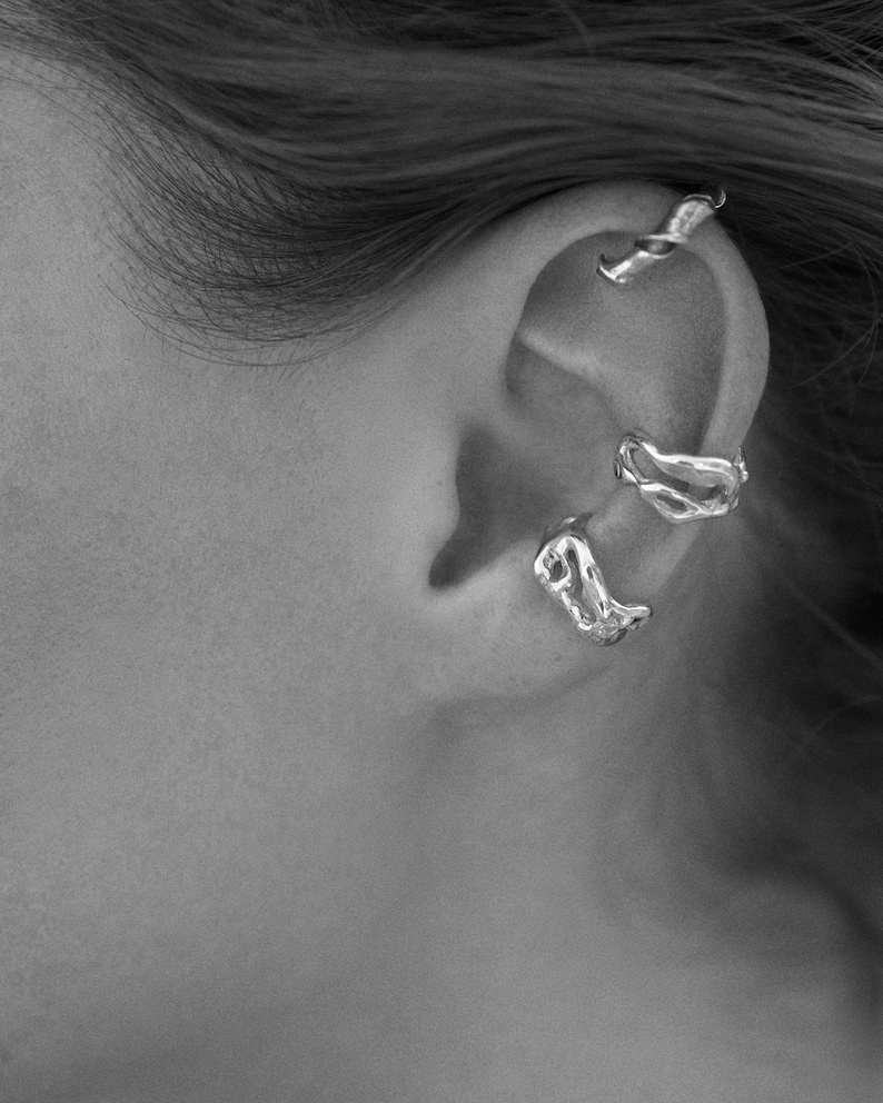 Pu&ograve; includere: Primo piano di un orecchio adornato da tre orecchini a polsino d'argento. Gli orecchini a polsino hanno un design organico e fluido, con un aspetto di metallo fuso strutturato. I gioielli sono ambientati sullo sfondo della pelle e dei capelli della persona.