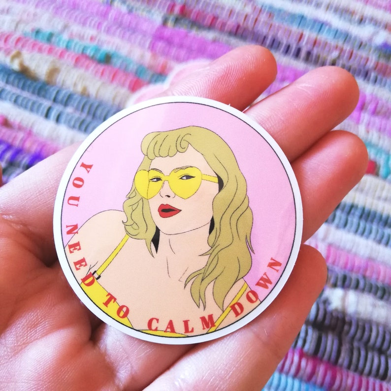 May include: A pink and white round sticker with a cartoon illustration of a woman with blonde hair wearing yellow heart-shaped sunglasses. The text "YOU NEED TO CALM DOWN" is written in red around the edge of the sticker.