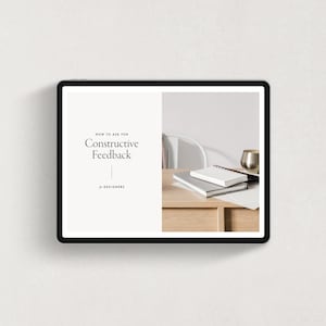 May include: A tablet screen displaying a white background with the text "How to Ask for Constructive Feedback for Designers". The right side of the screen shows a wooden desk with a white notebook, a silver pen, and a gold cup.