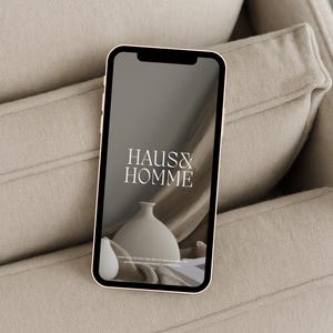 Könnte beinhalten: Ein Smartphone, auf dem eine Website mit dem Text "HAUS & HOMME" in Weiß auf schwarzem Hintergrund angezeigt wird. Die Website zeigt eine weiße Keramikvase auf einem beigefarbenen Stoffhintergrund.