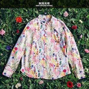 May include: A white long-sleeved shirt with a floral print featuring a variety of colors, including pink, yellow, blue, and green. The shirt is laid out on a bed of green leaves and flowers. The text "LIBERTY PRINT" is at the top of the image, and the text "S.M.L.の実物大パターン2枚" is at the bottom of the image.