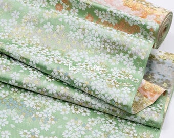 May include: A roll of light green fabric featuring a dense pattern of white cherry blossoms and gold metallic accents. The textile displays a repeating floral motif with subtle textured details and a soft sheen, showcasing a continuous design.