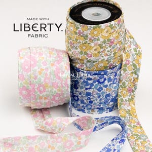 Può includere: Tre rotoli di nastro sbieco con motivi floreali in rosa, giallo e blu. Il testo MADE WITH LIBERTY FABRIC è visibile. Il nastro in tessuto è avvolto su bobine, con un'estremità di ogni rotolo che si estende per mostrare il bordo piegato.