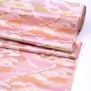 Puede incluir: Tela rosa con un patrón repetido de nubes, grullas y elementos florales. La tela tiene un brillo satinado y un borde enrollado. El texto "Forest Fabric" es visible en la tela.