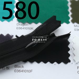 May include: Close-up of a black zipper with a black pull tab. The number "580" is printed in large black font above the zipper. The text "Dragon" and "Black" are printed on white tags. The website "www.forestfabric.com" is visible.