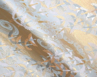 May include: White fabric with a gold and silver crane pattern. The cranes are flying in a flock and are surrounded by clouds. The fabric has a subtle sheen and a soft texture.