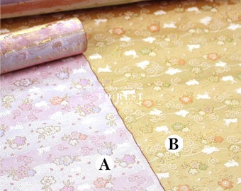 May include: Close-up of two patterned fabrics. One is pink with white stripes and floral designs, the other is gold with floral and cloud patterns. A rolled-up fabric with gold accents is visible.