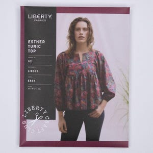 Pode incluir: Um envelope de molde de costura para a túnica Esther Tunic Top da Liberty Fabrics, número de padrão LIB301. A capa apresenta uma fotografia de uma pessoa vestindo uma túnica de manga comprida com padrão floral, decote fendido e cintura franzida.