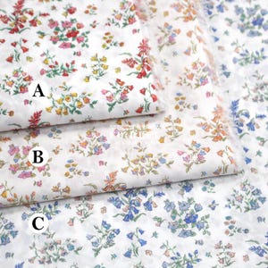 Pode incluir: Close-up de três amostras de tecido branco com padrões florais. Os padrões apresentam pequenas flores em tons de vermelho, rosa, amarelo, laranja e azul. O tecido tem uma borda delicada e ligeiramente desfiada, sugerindo uma textura macia.