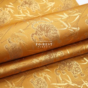 Può includere: Tessuto dorato con un motivo ripetuto di tigri e bambù. Il tessuto ha una lucentezza sottile e una sensazione di lusso.