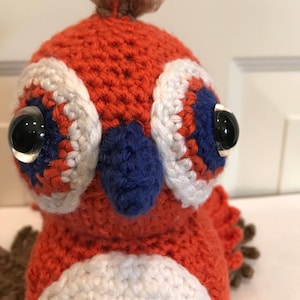 May include: A crocheted stuffed toy bird with orange, white, and blue details. The bird has a brown yarn tuft on its head and black button eyes.