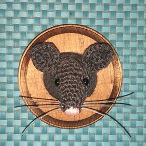 May include: A crocheted mouse head mounted in a wooden circle. The mouse is gray with black eyes and a pink nose. The mouse has long whiskers.