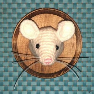 May include: A handcrafted, white crocheted mouse head mounted on a round wooden plaque. The mouse has black eyes, a pink nose, and long whiskers. The background is a teal woven pattern.